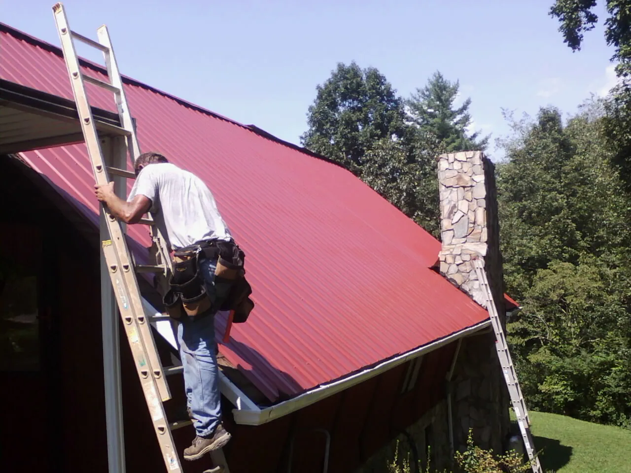 Roofer on a ladder beside a red metal roof and stone chimney — Soffit & Fascia Repair by Roofing Brothers in Rohnert Park